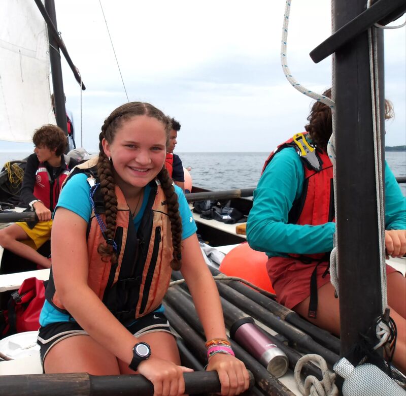 The photo shows three people on a sailboat. A young woman in the foreground is smiling and wearing a life jacket. Another person is behind her to the right, also wearing a life jacket and looking towards the front. A third person is partially visible in the background. The sailboat has a white sail and is on the water. The sky is overcast.